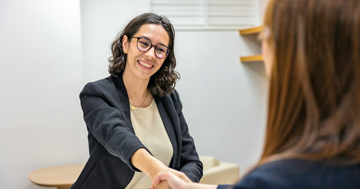 Lächelnde Frau in schwarzem Blazer schüttelt Hand einer anderen Person, die nur im Anschnitt von hinten zu sehen ist