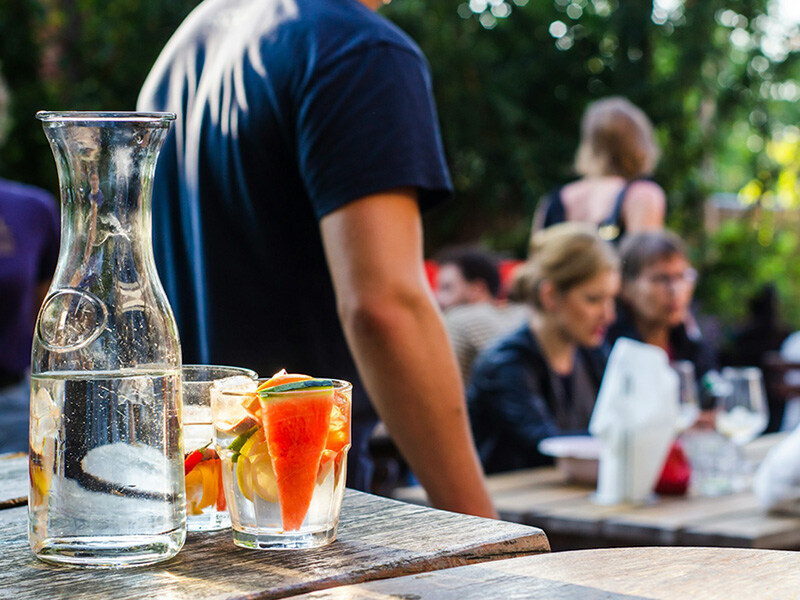 Ein Tisch im Freien mit einem Glas und einer Karaffe mit erfrischendem Wasser, im Hintergrund Gespräche führende Menschen bei einem Teamevent.