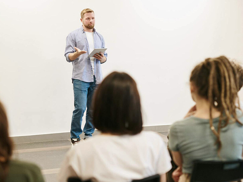 Ein Mann mit Tablet in der Hand präsentiert vor einer Gruppe sitzender Zuhörer, ein Beispiel für berufliche Weiterbildung und Mitarbeiterentwicklung.