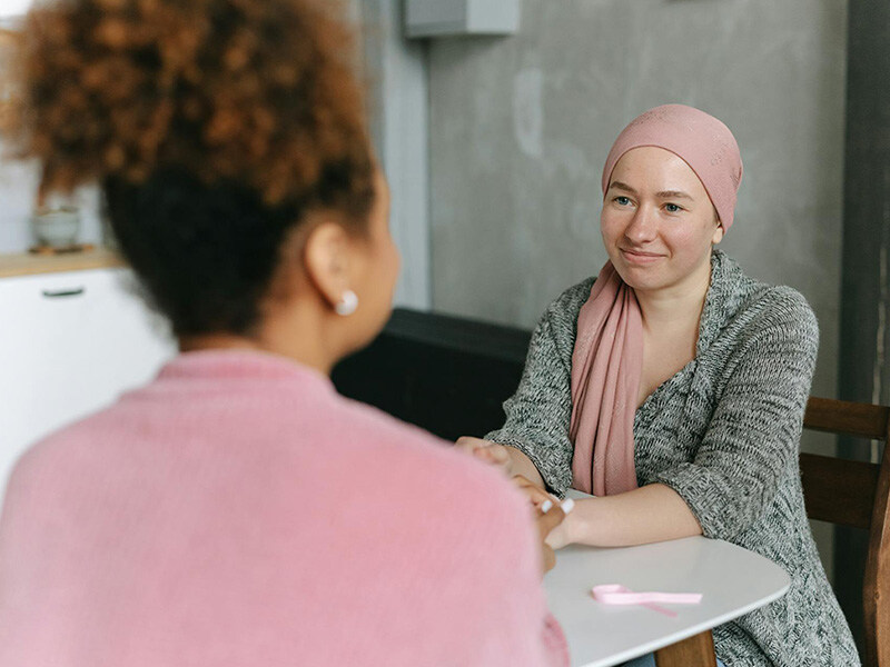 Zwei Frauen in einem beruhigenden Gespräch, eine mit Kopftuch sitzt zuversichtlich lächelnd gegenüber, ein Moment der Fürsorge in der Onkologiepflege.